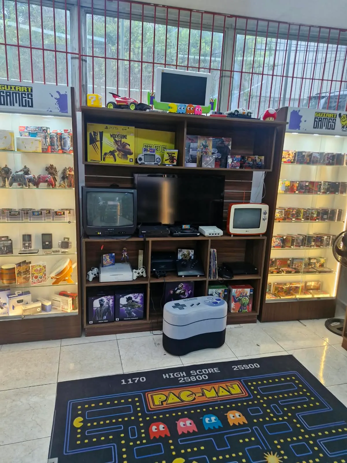 Interior da Guiart Games com TV de tubo, consoles e tapete do Pac-Man