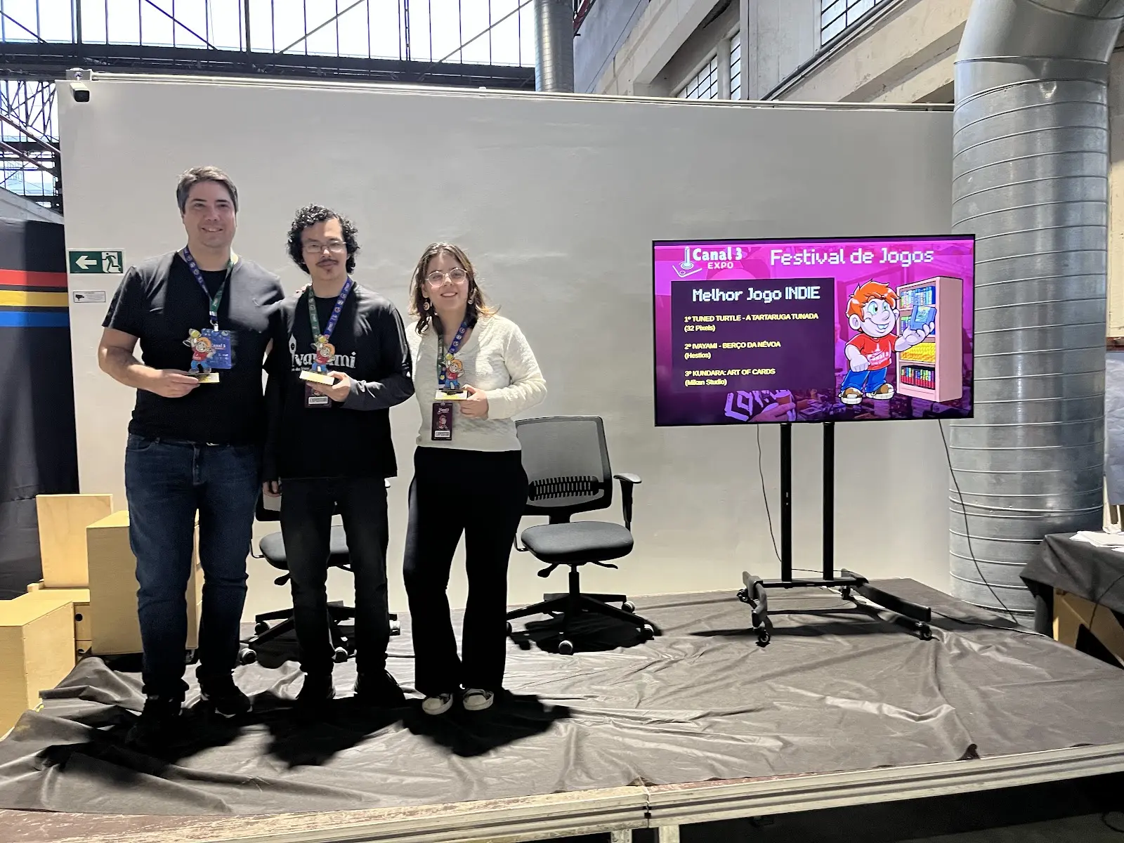 Vencedores do Melhor Jogo INDIE de 2025 no palco com troféus e o telão do Festival de Jogos